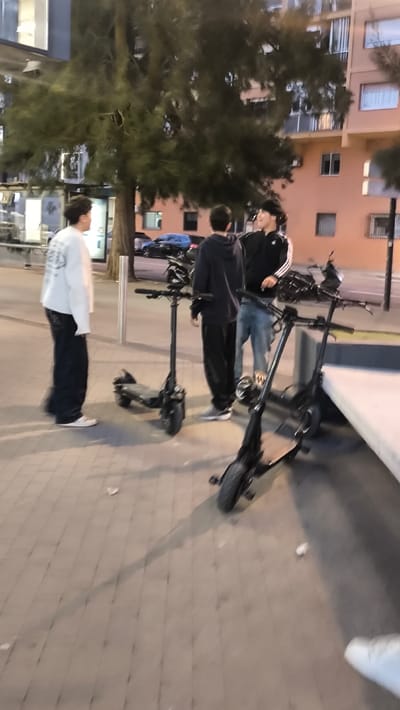 The image displays an outdoor urban scene in Ripollet, Spain, under artificial illumination, suggesting evening or nighttime. Three young male individuals are present on a paved sidewalk.

The individual on the left, partially blurred, wears a white long-sleeved shirt with a dark graphic and dark, wide-leg pants, facing towards the center-right. A black electric scooter is positioned directly in front of him. The middle individual is viewed from behind, wearing a dark hooded sweatshirt and dark pants, facing the third individual on the right. Another black electric scooter stands between the middle and right individuals. The third individual, on the right, wears a dark track jacket with white stripes on the sleeves and light-colored jeans; he is facing towards the center-left. All three individuals appear to be standing and interacting. A third black electric scooter is partially visible on the far right.

The ground consists of light-colored rectangular pavers. A silver bollard is situated between the left individual and the leftmost scooter. A light-colored concrete ledge or bench is present along the right edge of the frame. In the background, a street is visible with several parked vehicles, including a dark blue car. Beyond the street, multi-story residential buildings, one with a distinct peach hue, are discernible. A large tree with dark green foliage stands centrally, partially obscuring the background structures. Shadows are cast on the paved surface from the individuals and objects.