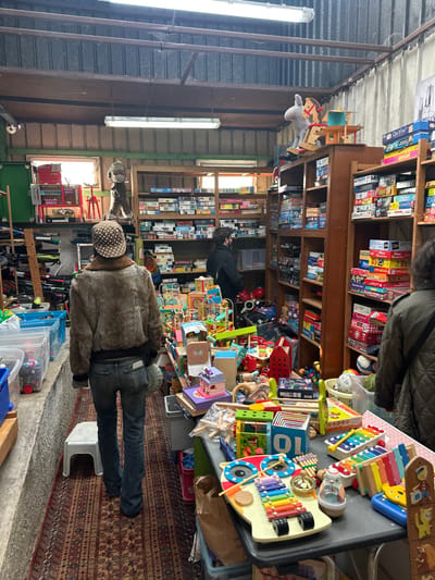 The image depicts an indoor scene within a second-hand store or market stall located in Neuilly-Plaisance, France. The environment is characterized by a cluttered display of various items, predominantly board games and children's toys.
The ceiling features exposed corrugated metal with visible wooden support beams and two suspended fluorescent strip lights. Natural light enters through two windows on the left wall, framed by green trim. The flooring consists of a patterned oriental-style rug covering a central aisle, with a concrete or rough surface visible along the left side.
Three individuals are present. In the foreground, a person is viewed from behind, walking away from the camera down the rug-covered aisle. This individual wears a brown faux fur bomber jacket, dark blue jeans, and a brown cap with white polka dots, carrying a light-colored pouch on their right hip. A small white plastic stool rests near their left foot on the rug.
In the midground, a second person is viewed from behind, wearing a dark jacket and a dark beanie or cap, browsing a tall wooden bookshelf on the right. Further right, partially visible, a third person's right shoulder and back are seen, wearing a dark quilted jacket, also appearing to browse merchandise.
The store's right side is dominated by multiple tall wooden bookshelves densely packed with various boxed board games. On top of one shelf, a gray plush rocking donkey with a red wooden base is prominently displayed. The midground and foreground feature a large rectangular table laden with numerous colorful children's toys, including wooden xylophones (one shaped like an owl), small dollhouses, building blocks, and assorted activity toys. Behind this table, more toys are visible, creating a dense arrangement. The left side of the store contains clear plastic storage bins, miscellaneous equipment, and an industrial-style cabinet. In the background, near the windows, a tall, light-brown fuzzy animal-like figure stands on a red base.