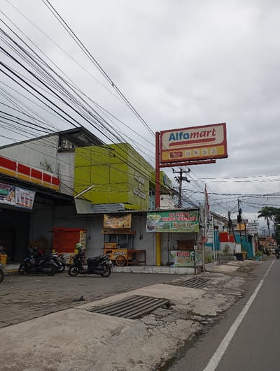 The scene is located at the coordinates -6.994 latitude and 107.555 longitude in Katapang, Indonesia. The image captures a street lined with buildings and shops. A prominent sign for "Alfamart," a convenience store chain, is visible on the left side of the street. The sign is mounted on a pole and is white with red and yellow lettering. The pole extends up into a cloudy sky and is encircled by a dense network of power lines. The buildings are two-story and have a mixture of brick and painted surfaces. The facade of the building behind the sign has a green and yellow paint scheme, while the building to the left is a single-story structure with a red, yellow, and white paint scheme. In front of the building, a group of three motorcycles are parked. They appear to be parked on a concrete slab, which slopes gently down to the street. There are additional shops and businesses along the street, including a "Tahu Goy" stand with a yellow and red sign. A sign for "Es Teller" can be seen on the right side of the street. The street is paved with asphalt and is bordered on each side by a curb and a walkway. The street is mostly empty, with only a few vehicles visible in the distance. The overall scene is one of a bustling commercial district.