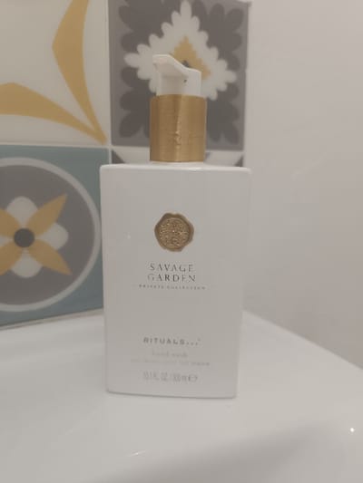 A white, rectangular bottle of "Savage Garden" hand wash by Rituals sits on a white surface. The bottle features a gold seal and a white pump with a gold base. Text on the bottle indicates it is part of a "Private Collection" and contains 10.1 fl oz / 300ml of hand wash. The bottle stands in front of a tiled background with geometric floral patterns in yellow, gray, and blue.