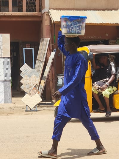 A street scene in Maiburiji, Nigeria, featuring a man in a blue outfit balancing a blue-lidded container on his head. Another person lies in a yellow motorized tricycle behind him. Ceramic tiles are displayed on a rack near a building with bars on the windows.