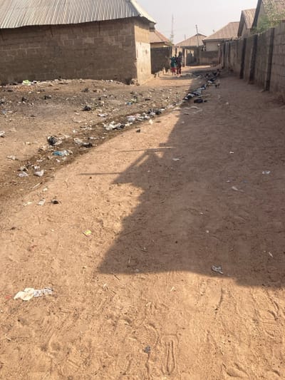 The image displays an unpaved dirt road traversing a residential area in Minna, Nigeria. The ground is dry, light brown earth, marked by numerous footprints. A prominent, elongated shadow, cast by an unseen object or structure, extends diagonally from the lower right foreground towards the mid-ground.

Along the left side of the road, a significant accumulation of refuse is present, consisting of various plastic items, paper, and unidentifiable debris. Multiple buildings constructed from exposed concrete blocks with corrugated metal roofs are visible on both sides of the road. Some structures on the left appear to be single-story and potentially unfinished. The right side of the path is bordered by a continuous wall built from concrete blocks.

In the mid-ground, three individuals are seen walking away from the camera along the dirt path. One person is distinctly visible wearing green clothing. Several chickens are observed on the path and near the refuse, foraging in the dirt. In the distant background, more residential structures are visible under a bright, slightly hazy sky, with a utility pole or tower present on the horizon.