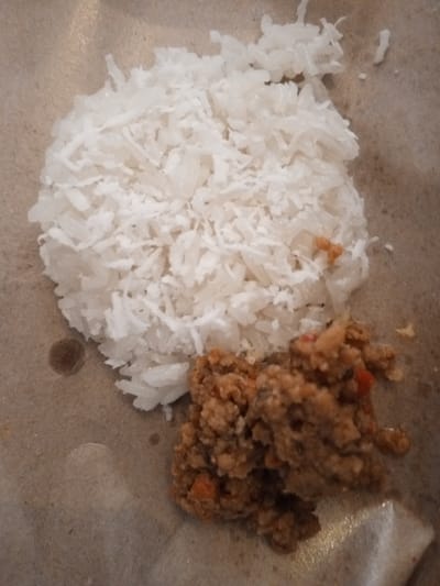 In Mvera Mission, Malawi, a close-up, overhead shot captures a meal served on a brown, textured surface, likely paper. The meal consists of a mound of fluffy white rice and a portion of a reddish-brown, minced food item, possibly meat or a stew with vegetables. The composition is simple, focusing solely on the food.
