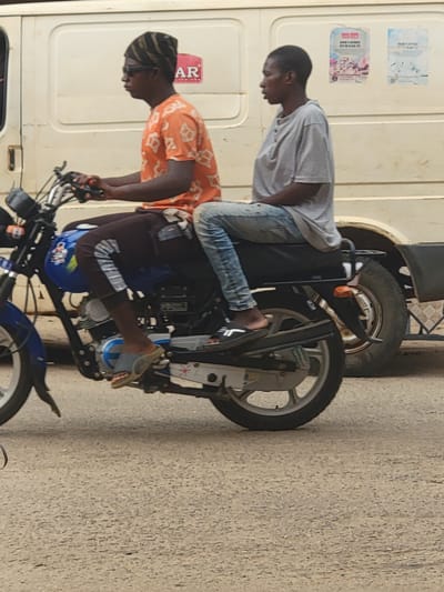 Two men are riding a motorcycle in what appears to be Maiburiji, Nigeria. One man in an orange shirt is driving the motorcycle. The passenger behind him is wearing a gray shirt and blue jeans. The motorcycle is blue and black. A white van is next to them. The road is made of asphalt.