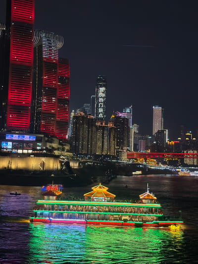 This nocturnal image captures a section of the cityscape in Nan'an District, China, viewed from an elevated perspective overlooking a wide river. In the immediate mid-ground, a large, multi-decked leisure boat, adorned with prominent green and red LED lighting along its hull and decks, navigates the dark water. The boat features traditional Chinese-style pagoda roofs, illuminated in golden-yellow, on its upper structures. Numerous individuals are visible on its open decks. To the left of this vessel, a smaller, dark rowboat or similar craft with a single occupant is present on the water.
The background consists of a dense urban environment dominated by illuminated skyscrapers and buildings. On the left, a cluster of modern high-rises stands out, with two exceptionally tall buildings brightly illuminated in solid red, featuring horizontal banding. One building further back displays a distinct white light pattern on its facade, resembling a starburst, and is topped with curved, illuminated structures. Along the riverbank, a fortified stone wall with an arched gateway is visible, illuminated by warm yellow lights, above which further buildings and structures are situated.
To the right of the central buildings, a complex array of skyscrapers and commercial structures glows with various white, yellow, and blue lights. A prominent red-lit bridge spans the river further in the background, connecting different parts of the city. The river's surface reflects the vivid lights from the boat and surrounding structures, creating patterns of green and red. The sky above is dark, with a faint, horizontal light streak visible in the upper right quadrant.
