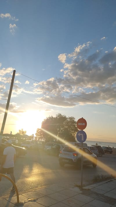 This vertical, candid photograph captures a street scene in Neoi Epivates, Greece, during sunset. The sun, low in the sky and casting a strong lens flare across the image, illuminates the scene with a warm, golden light. A tall, wooden utility pole stands on the left side of the frame, angled slightly.

In the foreground, a person wearing a white t-shirt and dark shorts is walking away from the camera, their back to the viewer. They are on a paved walkway with a checkered pattern, bordered by a yellow metal railing.

Further back, a line of parked cars is visible, including a white Dacia model with the license plate NN6 7764. A traffic sign with a red circle and a horizontal white bar, indicating "no entry" or "closed to all vehicles", is mounted above a blue circular sign with a white arrow pointing left.

Beyond the cars, a large tree with dense foliage stands, silhouetted against the bright sky. In the distance, the sea is visible, with a few small figures standing on the shore. The sky is a clear blue, with scattered white and gray clouds that are brightly lit by the setting sun. The overall atmosphere is serene, with a strong sense of the golden hour.