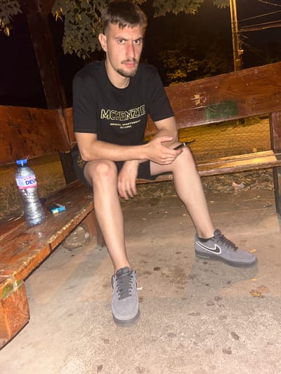A young adult male with short dark hair and a beard is sitting on a wooden bench in an outdoor setting at night. He is wearing a black t-shirt with a "MCKENZIE" logo and black shorts. The bench appears to be part of a covered structure with wooden supports and a thatched roof. A bottle of "DEVIN" mineral water and a pack of cigarettes are placed on the bench to his left. His left leg is extended forward, and his right leg is bent at the knee. He is holding a smartphone in his right hand. The ground is paved with concrete. In the background, there are trees and a utility pole with wires. The overall impression is a casual, possibly urban, nighttime scene. The location of Vratsa, Bulgaria is not discernible from the image.