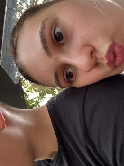 The media file is a close-up, upside-down selfie taken outdoors, likely in Gernsbach, Germany, given the context. Two individuals are partially visible. The foreground subject is a young person with fair skin, dark eyebrows, and brown eyes. They are making a puckered expression with their lips. Their hair, which appears light brown, is visible around their face. To the right, another individual's shoulder and neck are visible, wearing a dark t-shirt. The background shows dappled sunlight filtering through green foliage, suggesting it is daytime and likely fair weather. There is no visible text in the image. The scene appears casual and personal.