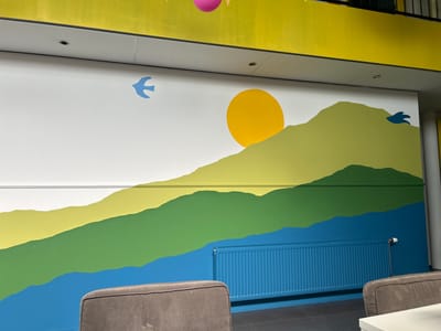 The image shows a mural on a white wall depicting a stylized landscape with rolling hills painted in shades of green and yellow, a blue body of water at the bottom, and a yellow sun. Two small blue birds are painted on the wall, one on the left and one on the right. A bright yellow band runs along the top of the wall. A blue radiator is mounted on the wall in front of the mural. Two upholstered chairs are partially visible in the foreground, and a white table is on the right edge of the image. The location is the reception area of a building in Lisse, Netherlands.