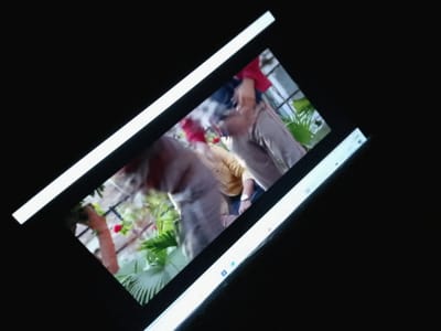 The image shows a close-up, angled shot of a laptop screen displaying a video. The video appears to be of people interacting outdoors in a garden or natural setting, with lush green foliage and a red flower visible. There are at least two people in the video, one wearing a red top and another in a yellow shirt. One person is reaching out, possibly holding something red, suggesting an activity or a gift exchange. The scene is blurry due to motion or focus, but the overall impression is a casual, outdoor gathering. The laptop screen itself is partially visible, with a white border along the top and bottom edges, and a sliver of the operating system interface at the bottom, showing icons that resemble Windows applications. The background of the image is black, indicating the surrounding environment where the laptop is being viewed, likely indoors or in low light conditions. There is no visible text within the video content. The location context provided is Zongoma Kyari, Nigeria.