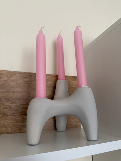 A close-up, eye-level, indoor shot captures a light gray, abstract-shaped concrete candleholder with three pink, tapered candles inserted into it. The candleholder has a unique, organic form with rounded legs and a central structure that holds the candles. The background consists of a textured wooden panel with a light beige wall above it. The scene is set on a white shelf. The image is well-lit, with soft shadows, and appears to be taken in a domestic environment. There are no people present in the image. The location of Pazardzhik, Bulgaria is not discernible from the visual information provided.