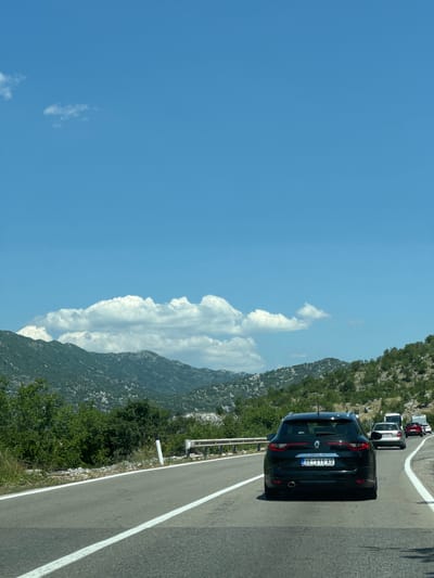 The image depicts a view from a vehicle traveling on a highway in the mountainous region of Cerovo, Montenegro. The road curves to the right, with a white line marking the lane division. A dark-colored Renault Megane station wagon, bearing the license plate "UE 215 AD," is the primary subject in the foreground, traveling in the same direction. Several other vehicles are visible ahead, including a white van, a silver sedan, and a red SUV. The landscape consists of lush green mountains covered in trees and vegetation, with rocky outcrops visible. The sky is a clear blue with scattered white clouds. A metal guardrail lines the left side of the road, separating it from the steep, vegetated embankment.