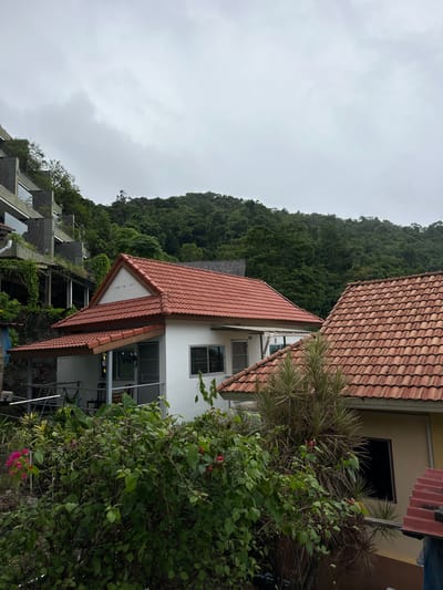 This media file shows a view of buildings and a lush green hillside, likely in Karon, Thailand. The scene is dominated by two houses with terracotta tile roofs. The foreground features dense green foliage, including flowering plants with pink blossoms. The houses appear to be built on a slope, with a terraced structure of concrete and glass visible on the left. The sky is overcast, suggesting a cloudy or possibly rainy day, common in tropical locations. There is no visible text in the image. The overall impression is one of a tropical, possibly humid, environment with residential structures nestled into the natural landscape.