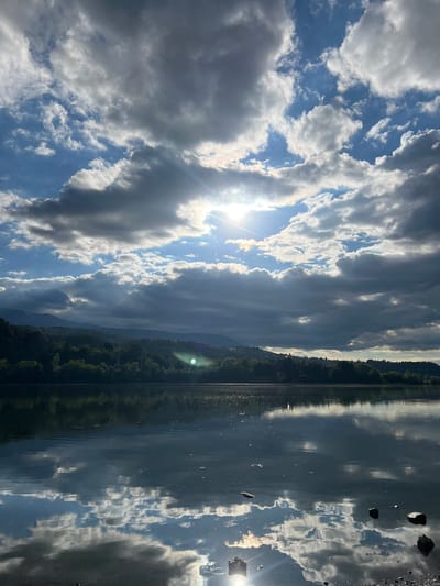 The image captures a serene lake scene in Pancharevo, Bulgaria, under a dramatic sky. The sun, partially obscured by a bank of dark clouds, casts bright rays and a noticeable lens flare onto the water. The sky is a dynamic mix of bright blue patches and heavy, textured clouds, some of which appear dark and foreboding while others are illuminated from behind. A treeline with lush green foliage borders the far side of the lake, backed by rolling hills and mountains that recede into the distance. The calm surface of the lake acts as a mirror, reflecting the sky, the treeline, and the distant mountains with remarkable clarity. Scattered rocks are visible along the foreground shoreline, and a few small, dark objects float on the water's surface. The overall atmosphere is one of natural beauty and tranquility, with the interplay of light and shadow creating a sense of depth and drama.