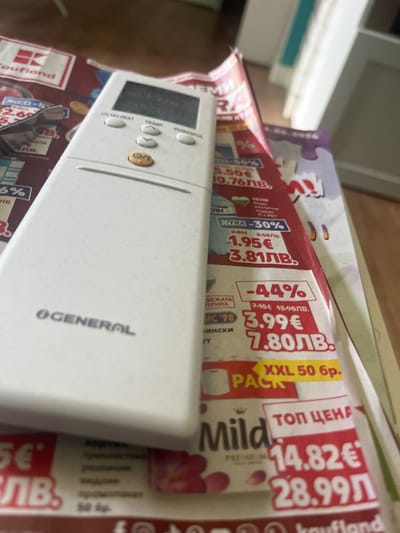 This image depicts a white 'GENERAL' brand air conditioner remote control resting on several open advertising flyers within an interior setting in Pokrovnik, Bulgaria. The remote features a blank LCD screen, a yellow/orange power button, and additional buttons labeled 'O/I HEAT', 'TEMP' (with up/down arrows), and 'POWERFUL'. Minor dust or smudges are visible on the remote's surface and screen. The underlying flyers display content primarily in Bulgarian, including prices in both Euros (€) and Bulgarian Lev (ЛВ.), and feature logos such as 'Kaufland'. Visible offers include discounts like '-30%' and '-44%', with original and discounted prices (e.g., 1.95€/3.81ЛВ., 3.99€/7.80ЛВ.) and product descriptions like 'XXL 50 бр.' and 'ТОП ЦЕНА' (Top Price) for items such as 'Mild'. The background behind the flyers is out of focus, showing light-colored walls and architectural elements, possibly a door frame, with a faint turquoise accent.