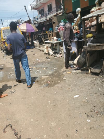 The image depicts a street scene in Shangisha, Nigeria. In the foreground, a young man wearing a dark blue t-shirt and blue jeans stands with his back to the camera, facing away from the viewer. To his right, an older man in a dark long-sleeved shirt and dark trousers stands facing a display of mannequins. The environment is characterized by a dusty, unpaved street littered with debris, including plastic bags, paper, and fabric scraps.
Behind the young man, a yellow commercial vehicle, likely a bus, is parked. A purple and tan umbrella is open near the bus. In the background, buildings with multiple stories line the street, some appearing to be shops. A white table with sewing machines is visible on the left side of the frame. The overall impression is of a bustling, somewhat unkempt market area. The lighting suggests it is daytime.