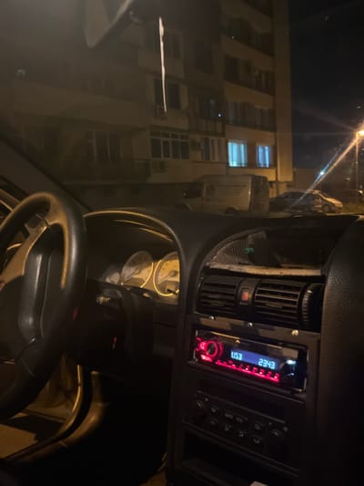 The image is a nighttime interior view of a car, likely parked on a street in Sliven, Bulgaria, with an apartment building visible in the background. The car's dashboard is in focus, showing the steering wheel, instrument cluster with illuminated gauges, and the car stereo. The stereo display shows "USB 2343" in blue text with red illumination. Below the stereo, there are controls for climate or audio functions. The exterior scene, visible through the car's windshield, includes streetlights, parked vehicles, and the lit windows of the apartment building, suggesting an urban residential area. The overall lighting is dim, indicative of nighttime conditions. There are no people visible in the image.