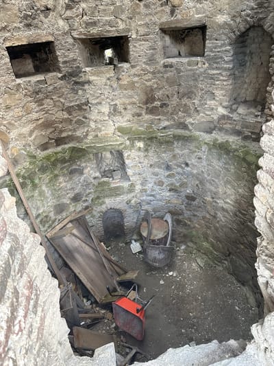 The image shows the interior of a partially ruined stone structure, likely a historical building or fortification, located in Shalikiantkari, Georgia. The walls are constructed from rough-hewn stones of varying sizes, some appearing weathered and covered in moss or lichen, indicating age and exposure to the elements. The interior is circular, with the stone walls curving inwards.
At the upper level of the structure, there are several square-shaped openings or embrasures built into the stone wall. These openings are dark and appear to lead to external spaces or be part of the building's defensive features.
The ground level of the interior is uneven and covered with dirt, gravel, and debris, suggesting a state of disrepair or ongoing excavation. Several objects are scattered on the ground. These include:
* A bright orange wheelbarrow, positioned near the center-left of the image.
* Two large, metal buckets or containers, one appearing to be made of metal with a rusted patina, located in the middle ground to the right.
* Several wooden planks and pieces of lumber, some leaning against the walls or piled on the ground.
* Other unidentifiable debris, including smaller stones and fragments of building materials.
There are no people visible in the image. The overall impression is of an ancient, weathered structure in a state of decay, possibly undergoing archaeological investigation or preservation work. The environment is enclosed and appears to be an interior space within the ruins.