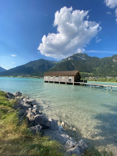 The image depicts a scenic view of a boathouse on a bright, clear day at Lärchenwiese, Austria. The boathouse, constructed of wood, stands on stilts over the turquoise water of a lake. A wooden pier extends from the boathouse into the lake. In the background, a range of tree-covered mountains with a prominent peak dominates the landscape, under a vibrant blue sky dotted with fluffy white clouds. The foreground features a rocky shoreline with green grass and vegetation. The water is shallow and transparent near the shore, revealing the rocky bottom. The scene is serene and untouched, with no people or animals clearly visible. The lighting suggests it is late morning or early afternoon.