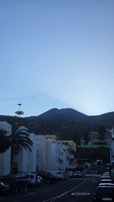 This image depicts a street scene in Guía de Isora, Spain, likely during twilight or early evening. The perspective is from an elevated position, looking down onto a street lined with cars parked along the side. Multiple apartment buildings of varying heights and architectural styles are visible on the left side of the street, with some featuring balconies and light-colored facades.
A prominent feature is the dark, silhouetted mountain range that dominates the background, with a faint glow along its peak suggesting either the setting sun or distant lights. In the foreground, a palm tree adds a tropical element to the scene. Power lines stretch across the sky, with a few birds perched on them. The street itself is paved and marked with white lines and circular road signs. Several cars are parked along the road, and one vehicle, a white van, appears to be parked slightly askew.
The time of day is indicated by the soft, diffused light and the deepening shadows, suggesting either dawn or dusk. The sky is a clear, pale blue. There are no people prominently visible in the image. The overall mood is quiet and peaceful, capturing a moment of transition between day and night in a residential area. A license plate on one of the cars is partially visible, showing "3502 BRP".