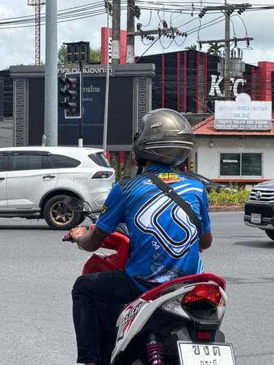 The image shows a man riding a scooter in Kathu, Thailand. The man is wearing a blue shirt with a racing-themed design and a silver helmet. He is in the middle of a road, with a white SUV to his left and a black Toyota SUV to his right. In the background, there is a KFC restaurant, traffic lights, and a traffic police box. The traffic lights are red, indicating that the man should stop. The road appears to be paved, and there is a sidewalk on the right side of the image. The sky is overcast.