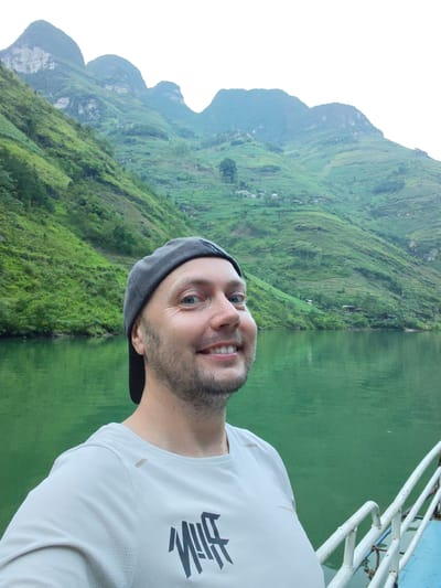 A fair-skinned man wearing a gray baseball cap and a light-colored t-shirt is smiling at the camera. He has blue eyes and a short beard. He is standing on what appears to be a boat, indicated by the white railing in the lower right corner of the frame. Behind him is a large body of green water. The background reveals steep, green, mountain slopes, suggesting a remote location. The image suggests a location in or near Đồng Văn Commune, Vietnam, characterized by steep, green-covered mountains and bodies of water. The man is taking a selfie.