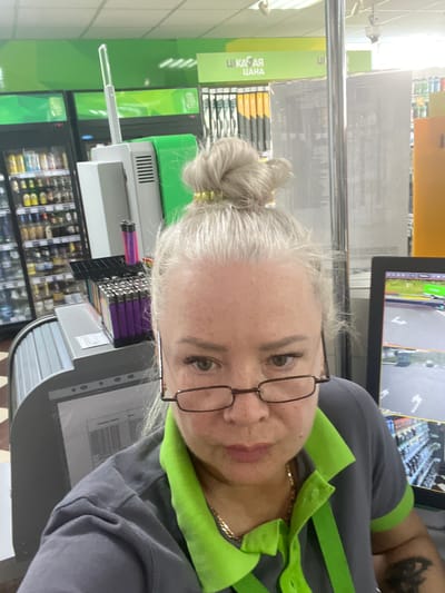 A woman with grey hair pulled into a bun is shown from the chest up, looking towards the camera. She is wearing a grey uniform with a lime green collar and trim, and has on glasses. Behind her, shelves stocked with goods and a computer monitor displaying a security camera feed are visible. A sign in the background reads "ЦІКАВАЯ ЦАНА" which translates to "INTERESTING PRICE". The scene appears to be inside a store, likely a supermarket or convenience store. The lighting suggests it could be daytime.