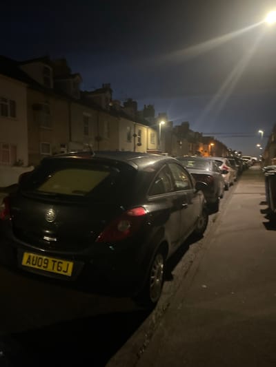A dark-filtered, rotated image shows a street scene in East Suffolk, United Kingdom, at night. The image is taken from a low angle looking up a narrow street lined with parked cars on the right and residential buildings on the left. The nearest vehicle, a dark hatchback, is partially in frame, displaying the license plate AU09 TGJ. Streetlights illuminate the scene unevenly. The buildings on the left appear to be multi-story townhouses. There is no indication of any people present.
