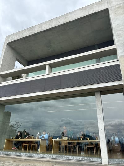 The image shows the exterior of a concrete building, seen from a low angle. The building is made of exposed concrete and has a series of large windows. The windows reflect a cloudy sky. The building is located at the Benesse House Museum in Naoshima, Japan. The coordinates provided place the image at the Benesse House Museum on Naoshima Island, Japan. Through the windows, a group of people can be seen seated at tables inside the museum, interacting and talking. They are seated in a row, facing the same direction. The people are wearing various casual clothes. The tables are made of wood and are arranged in a row along the windows. The floor appears to be made of light-colored stone. The image captures the museum's minimalist architecture and provides a glimpse into the daily life of visitors inside the building. The museum's interior features simple wooden furniture and windows that offer views of the cloudy sky.