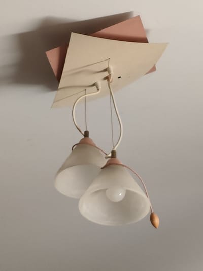 A ceiling lamp with two frosted glass shades hangs from the ceiling. The lamp has a tiered design with triangular elements in pink and beige. One shade has a visible lightbulb. A wooden pull-chain is attached to one of the shades. The scene is indoors, likely a residential interior, in Андрэеўшчынскі сельскі Савет, Belarus. The lighting is dim, casting shadows on the ceiling.