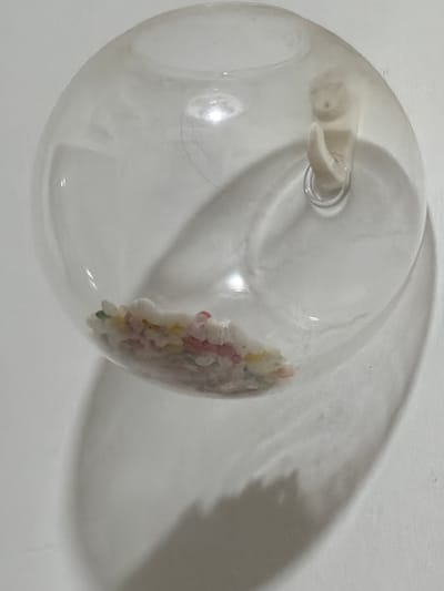 A clear, transparent spherical object rests on a plain, uniformly lit, light-colored surface. Within the lower portion of the sphere, a small cluster of irregularly shaped granules is visible, exhibiting various colors including white, pink, yellow, red, and hints of green. Near the upper right interior of the sphere, a light-beige, amorphous object with an organic, textured surface is present. A small, reflective metallic ring is attached to a hook-like protrusion extending downwards from this object. A distinct, elongated shadow cast by the spherical object extends downwards and to the right on the background surface. No people or actions are depicted. The immediate environment is stark, offering no discernible identifying features related to a specific city or district.