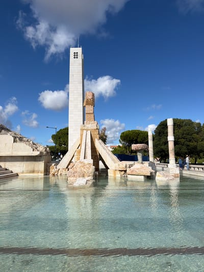 The image shows the Monument to the Discoveries in Lisbon, Portugal. The monument is a tall, white stone structure with a series of decorative windows near the top. It is partially reflected in the water in the foreground. To the right of the monument is a stone fountain-like structure with various levels and sections, also reflecting in the water. In the background, behind the fountain, are green trees and some columns or remnants of structures. The sky above is blue with scattered white clouds.