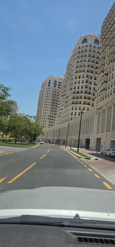 The image, captured from the perspective of someone inside a vehicle, showcases a sunlit street scene in Doha, Qatar, likely within a modern development characterized by grand architecture. Towering cream-colored buildings with numerous balconies and arched windows dominate the background, suggesting a residential or hotel complex. The street itself is paved with asphalt and marked with yellow lines, indicating lanes for traffic. Green trees line the left side of the road, with manicured lawns and decorative bollards bordering the pavement on the right. Several cars are visible, including a white sedan parked on the right and another white car driving in the distance. Traffic signs, including speed limits and directional arrows, are strategically placed along the roadside. The sky is a clear, vibrant blue, typical of the region's climate. The overall impression is one of order, spaciousness, and contemporary urban design.