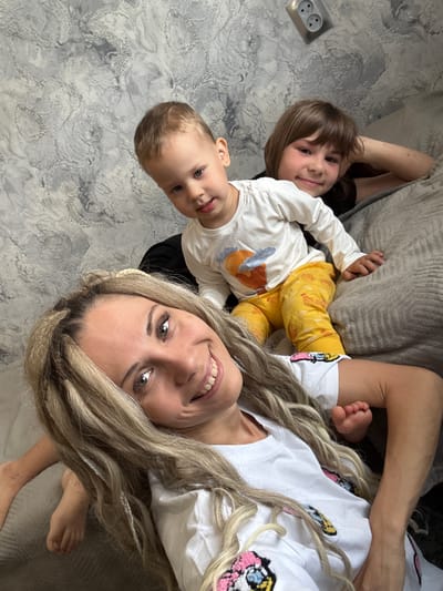 The image is a selfie taken from a high angle, depicting a woman and two children. The woman, with blonde hair, is smiling at the camera. A toddler, likely a boy, is positioned to her left. Another child, possibly slightly older, is behind the toddler. The setting appears to be indoors, possibly in a home. The presence of an electrical outlet on the wall suggests an interior setting. The location in the city of Rusanovo, Russia cannot be confirmed from this image.