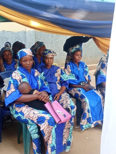 The scene, located in Doma, Nigeria, depicts an outdoor gathering under a temporary canopy. Seven individuals, primarily women, are visible, with five prominently seated. All observable women wear matching uniforms consisting of a blue gown adorned with blue, teal, and beige patterned fabric, and corresponding head coverings. In the foreground, a woman in this uniform sits on a blue plastic chair, holding a sleeping infant wrapped in dark fabric against her chest. A small pink bag with a white emblem rests on her lap. To her immediate right, another woman, similarly dressed but wearing a patterned cap, is seated, looking forward. Further right, a third woman in identical attire, including a headwrap and a small golden pendant, is also seated. Additional women in similar uniforms are partially visible in the background on the left and right. Overhead, blue and yellow fabric drapes form a canopy. The background features a light-colored concrete wall, and the ground surface appears to be dirt.