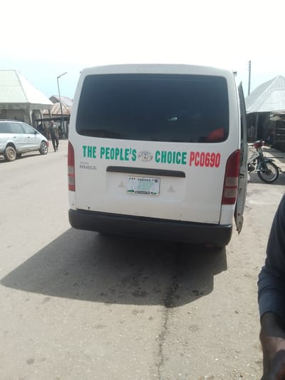 The image shows the rear of a white Toyota Hiace van parked on an asphalt road. The van has a dark tinted rear window and a partially opened rear passenger door. Green lettering on the back of the van reads "THE PEOPLE'S CHOICE" with the Toyota logo in between. To the right of "CHOICE," red lettering spells out "PC0690." A license plate is visible below the lettering, with "TARABA" and "268-901" printed on it. The license plate also includes the Toyota logo.
In the background, to the left of the van, a silver minivan is parked. A man in a green shirt is walking on the road between the two vehicles. Further in the background, there are several buildings with corrugated metal roofs, suggesting a marketplace or residential area. A motorcycle is parked to the right of the van, near one of the buildings. The sky is bright and appears to be daytime, with an overcast or hazy condition. The overall scene suggests a typical street in a Nigerian town.