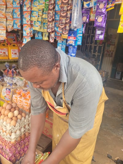 A man stands at a market stall in Madalla, Nigeria. He's light-skinned with a close-cropped, dark hair and wears a grey collared shirt and yellow covering. The stall displays a variety of goods including Cadbury chocolate drinks, Nasco cornflakes, oats, Kings cooking oil, Top 2 bread, eggs, and Maggi. He is attending to eggs at the stand.