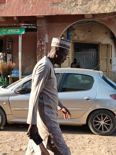 An adult male with dark skin walks left across an unpaved street in Gwagwarwa, Nigeria. He wears a light beige long-sleeved tunic and a white cap with dark geometric patterns. He holds a dark smartphone in his left hand, and his gaze is directed downwards and to the left. Behind the man, a silver five-door hatchback, consistent with a Peugeot 206, is parked parallel to the street. The car shows minor damage or dirt near the rocker panel and has multi-spoke dark rims on the visible rear wheel.
In the background, a weathered building features a reddish-brown upper facade with peeling paint and a lighter, off-white lower section. An arched doorway in the building reveals a dark interior, inside which two or three individuals are partially visible; one appears to be a standing dark-skinned male. A blue pipe runs vertically on the building's facade. To the far left, a green sign with white text, partially reading "ENERGY TO GO FURTHER," is mounted above a display of brown bundled objects (possibly brooms) in a black bucket, green plastic crates, and blue jerrycans. The ground is a light brown dirt surface. The scene is illuminated by strong daylight, casting shadows.