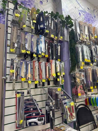 This image shows a wall display of kitchen knives and accessories in a store. The location is likely a department or specialty kitchen store in the city of Klin, Russia, given the presence of Russian text on the product packaging. There are various types of knives on display, including chef's knives, carving knives, and utility knives. Knife sharpeners, scissors, and a magnetic knife holder are also displayed. The products are arranged on metal racks against a white wall with green vertical stripes and some decorative accents such as greenery and purple flowers in the background. Small, yellow price tags are attached to the products. The scene appears to be a retail setting within a store offering kitchenware.