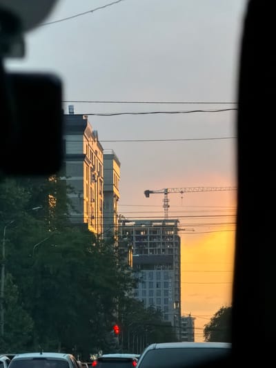 This media file captures a view of a cityscape during what appears to be sunset or sunrise, indicated by the warm, orange glow in the sky. The scene is dominated by modern architecture, with several buildings under construction. A prominent construction crane is visible against the vibrant sky, suggesting ongoing development. One of the buildings features large, block lettering that spells out "REGNUM". The overall setting is urban, likely in Bishkek, Kyrgyzstan, as indicated by the location context. Trees, possibly part of a park or lining a street, are also present, partially obscuring some of the buildings and adding a touch of nature to the concrete landscape. The image is framed by what seems to be the interior of a vehicle, with dark elements at the top and bottom, suggesting the photo was taken from a car window. The presence of traffic lights, one showing red, implies the photo was taken during traffic. The lighting suggests it's either early morning or late evening.