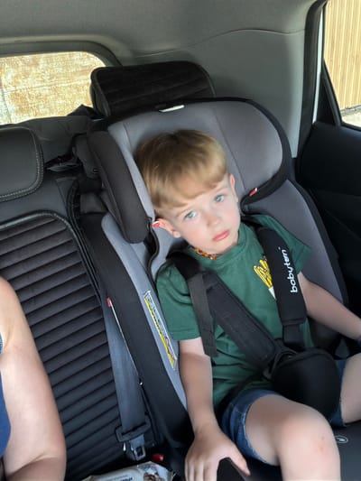 A young boy with blond hair and blue eyes lies on his back in a car seat. He is wearing a green t-shirt and denim shorts. The car seat is gray and black, with a "babyten" logo visible on the shoulder strap. The boy's head is tilted back, and he is looking up with a neutral expression. The setting appears to be the interior of a car, with a window visible in the background. The lighting suggests it is daytime. The text "babyten" is visible on a strap. The location context is Ust'-Dzhigutinka, Russia.