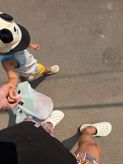 In Denasri Kulon, Indonesia, a low-angle, full-body shot captures a parent holding a child's hand while walking on a paved road. The child, facing away from the camera and to the left, wears a white sleeveless shirt with blue trim, yellow shorts, and cream-colored shoes. A stuffed panda-themed hat adorns the child's head. In the parent's hand, a transparent plastic bag filled with various items is visible. The parent is wearing black shorts with a colorful abstract pattern and white Crocs-style shoes. The ground is rough asphalt, showing subtle shadows from an unseen object, suggesting a sunny day. The scene is devoid of other people or vehicles, focusing solely on the parent-child interaction and their immediate surroundings.