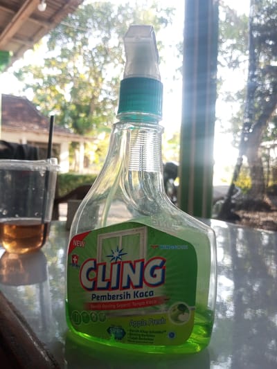 The image shows a clear plastic bottle of Cling window cleaner with a green liquid inside, positioned on a reflective surface. The bottle has a spray nozzle and a green label with the brand name "CLING" prominently displayed in red and blue letters. The label also features text in Indonesian indicating it is a glass cleaner. To the left of the bottle is a clear plastic cup with a brown liquid, possibly tea, and a black straw. In the background, out-of-focus, are trees and structures, suggesting an outdoor or semi-outdoor setting. The lighting indicates it is daytime, with sunlight filtering through the leaves. The presence of the "CLING" brand, a common household product, and the general appearance of the scene do not specifically indicate the location as Ketoyan, Indonesia, although the provided context suggests this is the case.