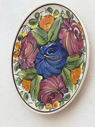 A round, decorative ceramic plate with a floral design is affixed to a white, textured wall. The plate features a central blue flower surrounded by large, stylized red and purple flowers, orange blossoms, and green leaves. A thin brown rim encircles the plate. The wall appears to be an exterior surface, possibly of a building in Torrevieja, Spain, given the context of similar imagery from the region. The lighting suggests daylight.