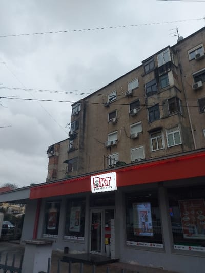 The image depicts an urban street scene in Tiranë, Albania, on what appears to be an overcast day. The frame is dominated by a multi-story apartment building on the left and a modern storefront on the right.
The apartment building, constructed with a light brown or beige facade, shows signs of age and features numerous windows across its multiple floors. Many of these windows have external air conditioning units attached, and several small balconies are also visible. An antenna is mounted on the building's roof, extending into the cloudy sky.
Adjacent to the apartment building, on the right side of the frame, is a contemporary storefront with a prominent red awning or fascia. A brightly lit, rectangular sign is affixed to the facade, displaying the large red letters "BKT." Below "BKT," smaller text is visible, which reads "Banka Kombëtare Tregtare," indicating "National Commercial Bank." The 'T' in BKT is stylized with a bird or eagle motif. The storefront features large glass display windows that showcase various advertisements and promotional materials, likely related to banking services. One visible advertisement in a window appears to mention "Vip club." The entrance to the bank is situated between the display windows.
The sky above is uniformly gray and heavily overcast, suggesting a cloudy day. Several utility wires crisscross the upper portion of the image. In the lower right corner, behind the bank structure, some hints of greenery, possibly trees or bushes, can be seen.
No people are visible, and no specific events or activities are taking place beyond the implied operations of the bank. The overall scene portrays a typical commercial and residential street environment in a city.