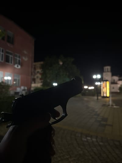 The image depicts an urban scene at night, likely in Kostinbrod, Bulgaria. In the foreground, a human hand holds a dark, pistol-shaped object, silhouetted against the illuminated background. The object possesses characteristics of a semi-automatic handgun, with the index finger positioned on or near the trigger. The background, which is out of focus, reveals a paved street or square. To the left, a multi-story building with a reddish-brown facade and numerous windows is visible. To the far right, a distinct white building featuring a prominent clock tower is discernible, identified as the Municipality of Kostinbrod. Multiple streetlights cast bright, unfocused light sources across the scene, and a vertically oriented, brightly lit sign with yellow and orange illumination is present in the mid-ground right. The sky is dark, indicating nighttime conditions. No other individuals are clearly visible.