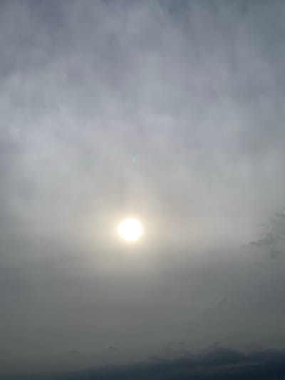 The image captures a hazy sky with the sun as a bright, diffused disc at the center. The sky is filled with clouds, appearing in varying shades of gray and white, suggesting an overcast or foggy atmosphere. The lower portion of the image shows darker, more opaque clouds, creating a layered effect. The overall scene is monochromatic, with the bright sun piercing through the dense cloud cover. There are no discernible human figures, objects, or specific landmarks visible in the image, making it impossible to pinpoint a specific location within Kuru Karama, Nigeria, based solely on the visual information provided. The environment depicted is solely the sky.