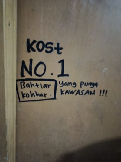 The image shows a portion of a wall with writing on it. The writing is in black marker. At the top, it says "kost" with a plus sign next to it. Below that, it says "NO. 1". To the left of "NO. 1" there is a handwritten box containing the words "Bahtlar kohhar .". To the right of "NO. 1", it says "yang punya KAWASAN !!!". The wall appears to be made of particle board or a similar material and has a wood-like texture. A vertical wooden element is visible on the left edge of the image. The overall lighting suggests an indoor setting, possibly a room or hallway. The writing appears to be a casual, handwritten label or sign, common in informal settings in Indonesia, such as boarding houses or small businesses. The specific location in Depok, Indonesia cannot be determined from the image alone, as the content is a generic sign.