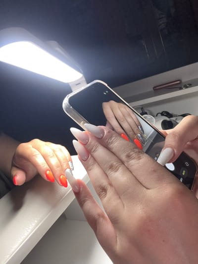In this close-up shot, a person's hands are prominently featured, showcasing elaborate, long, almond-shaped acrylic nails. One hand is holding a smartphone, angled to capture a reflection of the other hand and its nails. The nails on the reflected hand are a vibrant orange, with some accented with glittery detailing. The hand holding the phone has nails that are a shimmering white, with a subtle ombre effect transitioning to clear at the tips.
The scene appears to be indoors, possibly a home or a beauty salon, given the presence of a bright, rectangular light source in the lower left corner, likely used for photography or illumination. The background is dark and out of focus, with subtle hints of metallic objects on the left, possibly part of a setup or decor. The overall lighting suggests it could be daytime or evening, with the artificial light being the primary source.
The activity is the act of photographing one's own nails, a common practice for sharing beauty achievements online. The focus is entirely on the hands and the aesthetic of the nails, suggesting a personal interest in nail art or a promotion of nail services. There is no clear indication of emotion, but the deliberate framing and lighting imply an effort to present the nails in the best possible light.
Visible text on the phone screen includes Russian words: "ФОТО" (FOTO - Photo) and "0.5 1.3x", suggesting the camera settings or modes. The setting is given as Dmitrov, Russia.