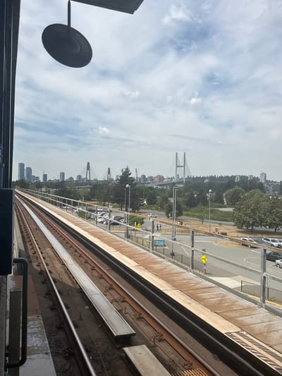 The image depicts a view from a Skytrain platform in Surrey, Canada, looking out towards the city skyline and a bridge. The foreground features Skytrain tracks, with the platform edge visible on the right. Beyond the tracks, a parking lot with several cars is situated, and further in the distance, a cityscape is visible with tall buildings and bridges spanning a body of water. Green trees and vegetation dot the landscape. The sky is overcast with scattered clouds. There are a few pedestrians visible on the sidewalk near the parking lot. A dark, circular light fixture hangs from the overhang of the Skytrain station in the upper left corner of the image.